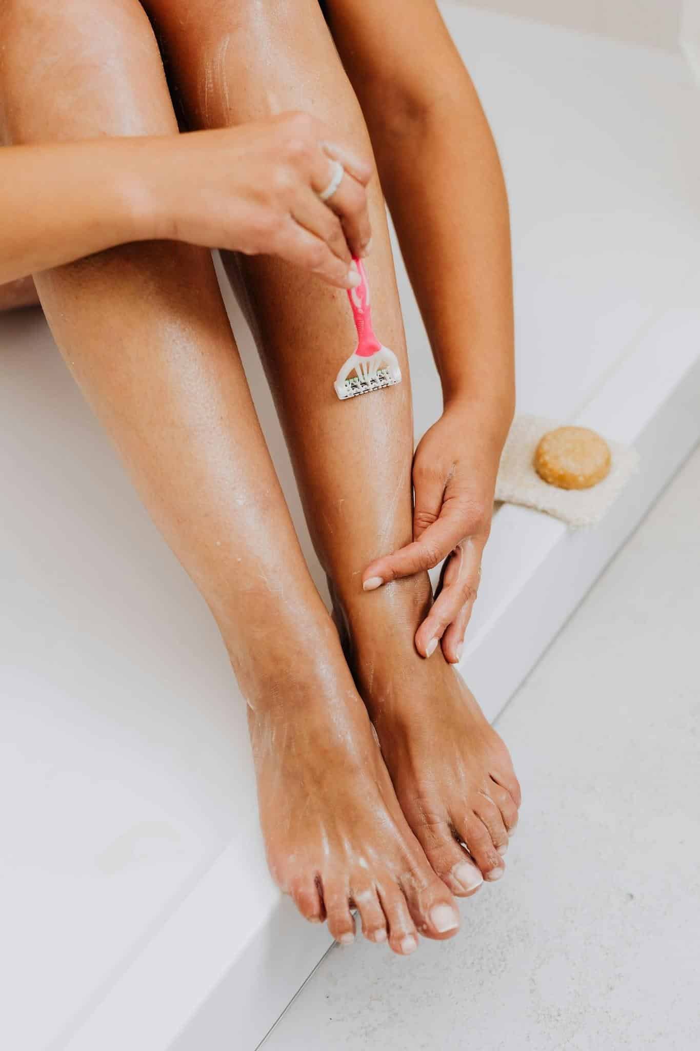 A person shaving their leg with a pink razor whilst sitting on the edge of a bath; a loofah and soap are nearby.