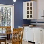 A kitchen with white cupboards, a dishwasher, and a round wooden dining table with four chairs near a large window and blue feature wall.