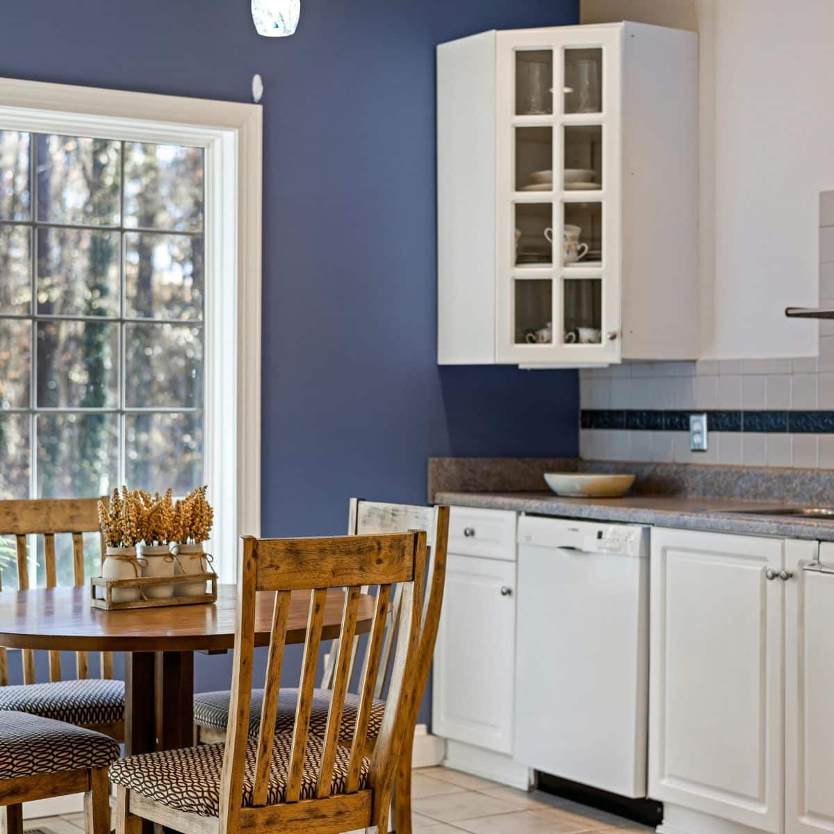 A kitchen with white cupboards, a dishwasher, and a round wooden dining table with four chairs near a large window and blue feature wall.