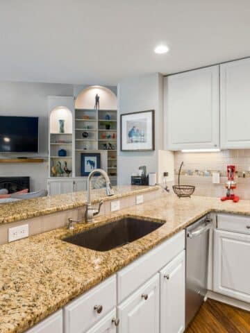 Modern kitchen with granite worktops, white cupboards, stainless steel sink, and a view into a living area with built-in shelves and a wall-mounted TV.