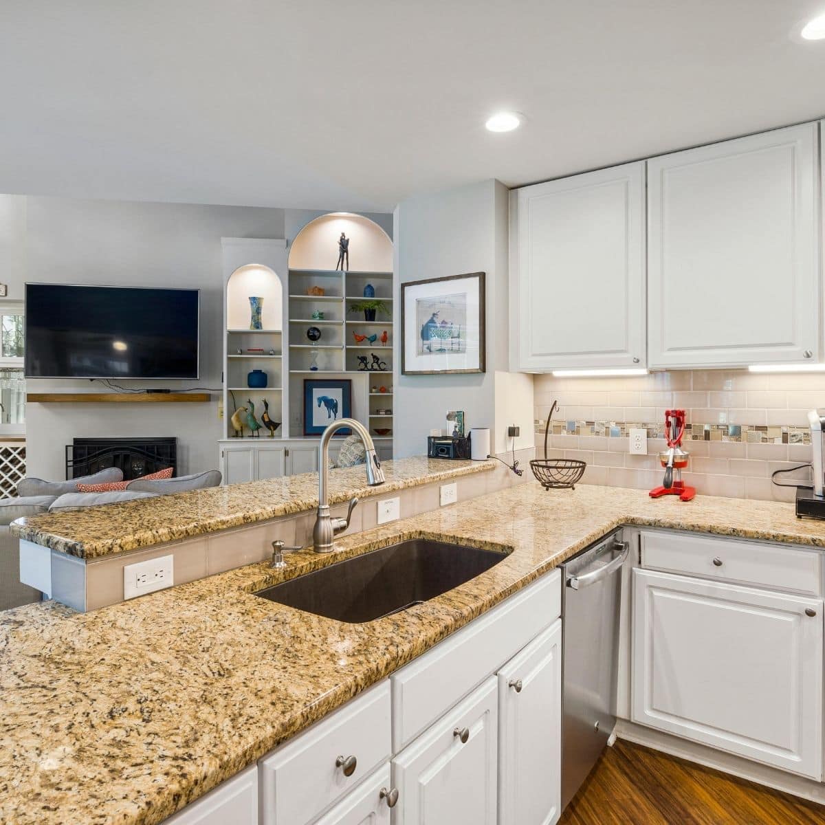 Modern kitchen with granite worktops, white cupboards, stainless steel sink, and a view into a living area with built-in shelves and a wall-mounted TV.