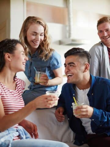 Four young adults sit and stand indoors, smiling and talking whilst holding drinks, suggesting a casual social gathering.