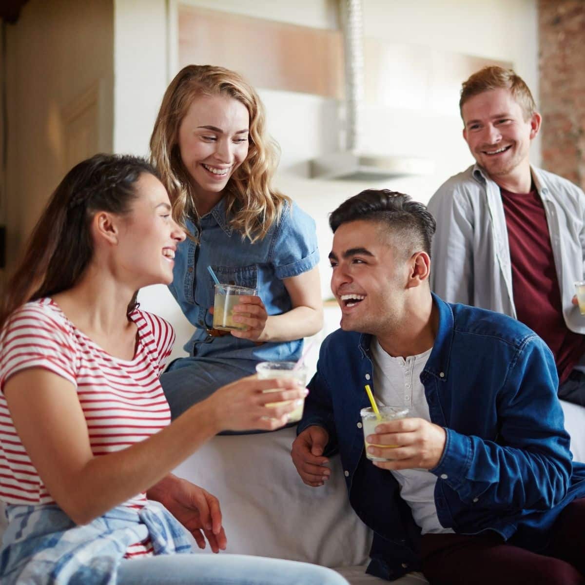 Four young adults sit and stand indoors, smiling and talking whilst holding drinks, suggesting a casual social gathering.