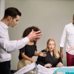 Four people in business attire are having a heated discussion around a desk with documents and folders, with one woman seated and others standing.