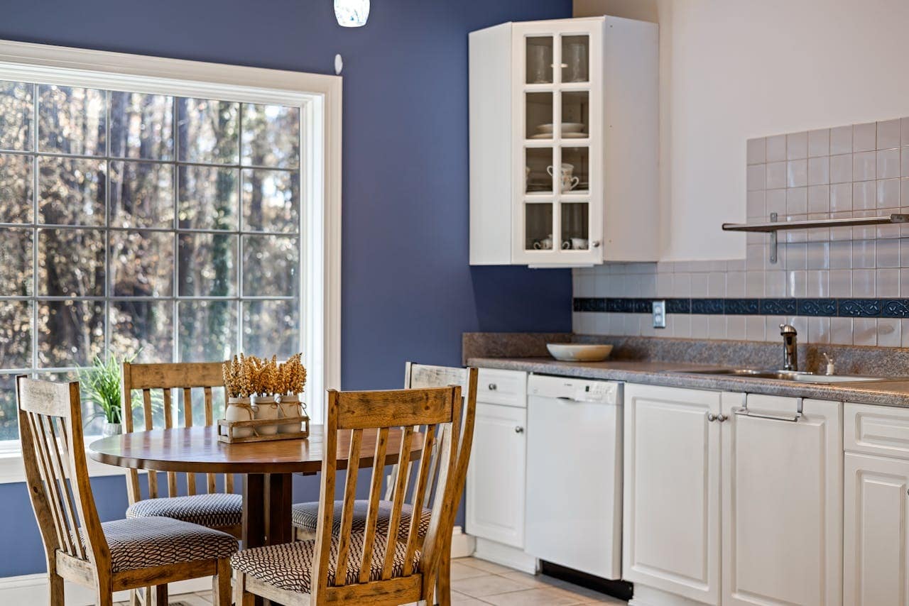 A kitchen with white cupboards, a round wooden dining table with four chairs, a large window, and a blue feature wall. Dried flowers decorate the table.