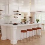 Modern kitchen with a large white island, three brown stools, open shelves, pendant lights, and a dining area with windows in the background.