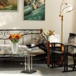 A cosy living room with a glass coffee table, a daybed with floral cushions, flowers in vases, a standard lamp, and framed art on the wall.