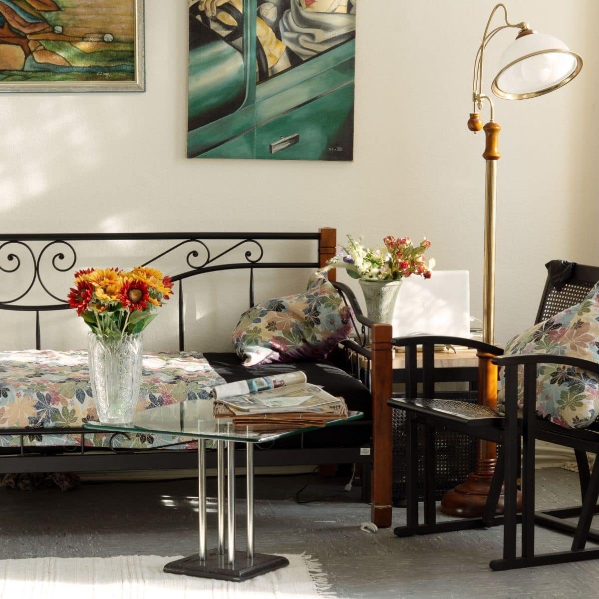 A cosy living room with a glass coffee table, a daybed with floral cushions, flowers in vases, a standard lamp, and framed art on the wall.