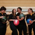 Four people in matching bowling uniforms stand together in a bowling alley, each holding a bowling ball and smiling.