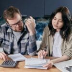 Two people sit at a table reviewing documents with a calculator and notebook, appearing focused and engaged in paperwork.