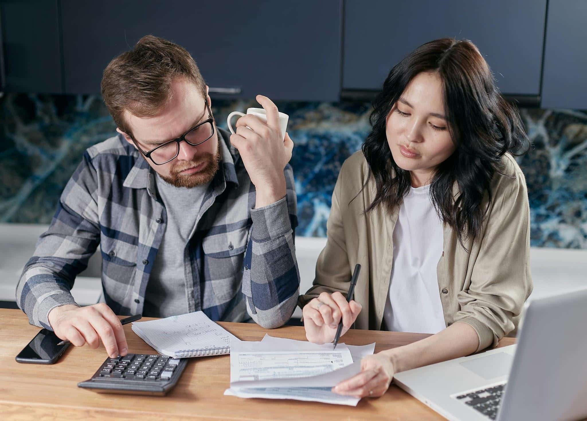 A man and woman sit at a table reviewing documents, using a calculator and writing notes, with a laptop and phone nearby.