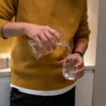 A person in a mustard jumper pours water from a clear jug into a glass in a kitchen setting.