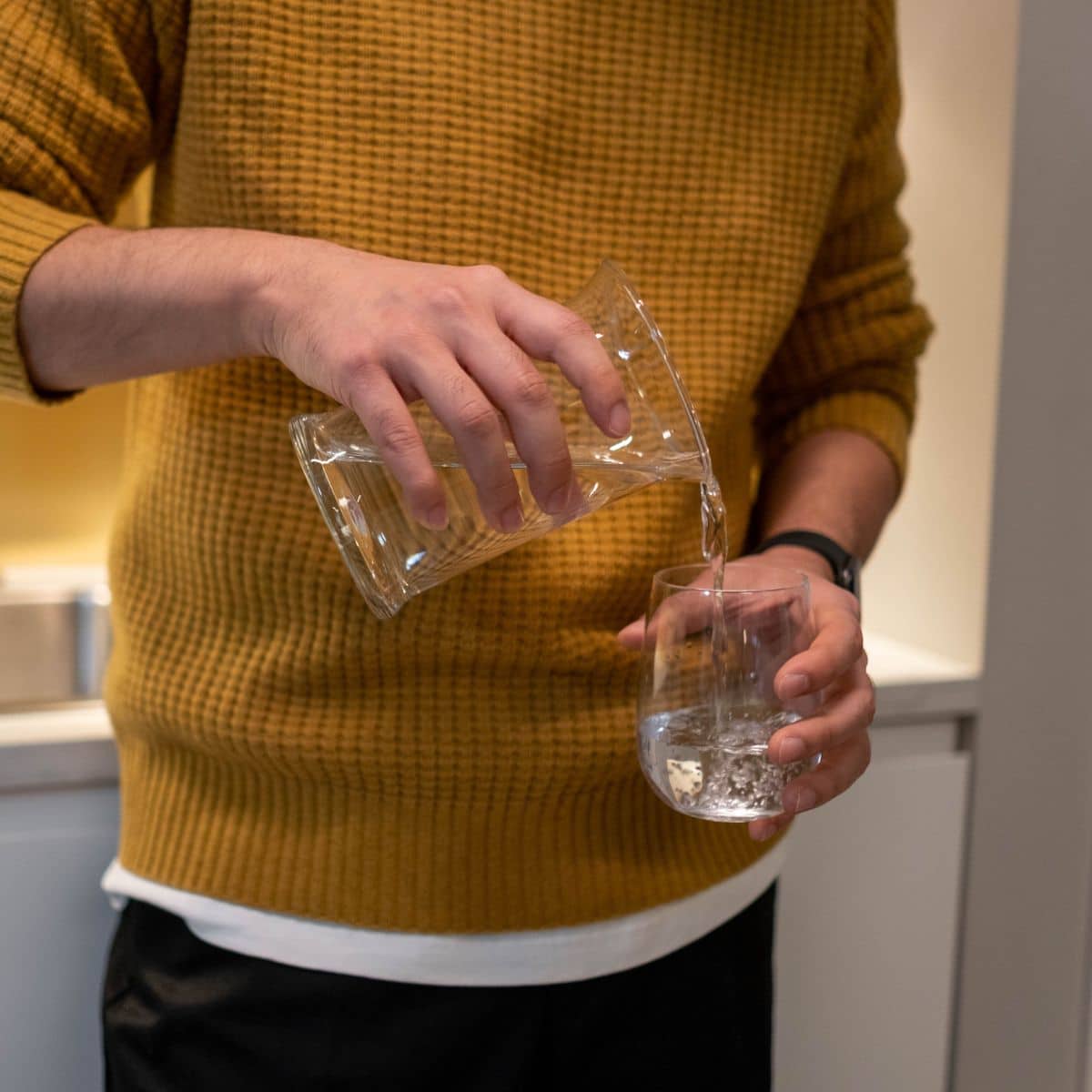 A person in a mustard jumper pours water from a clear jug into a glass in a kitchen setting.