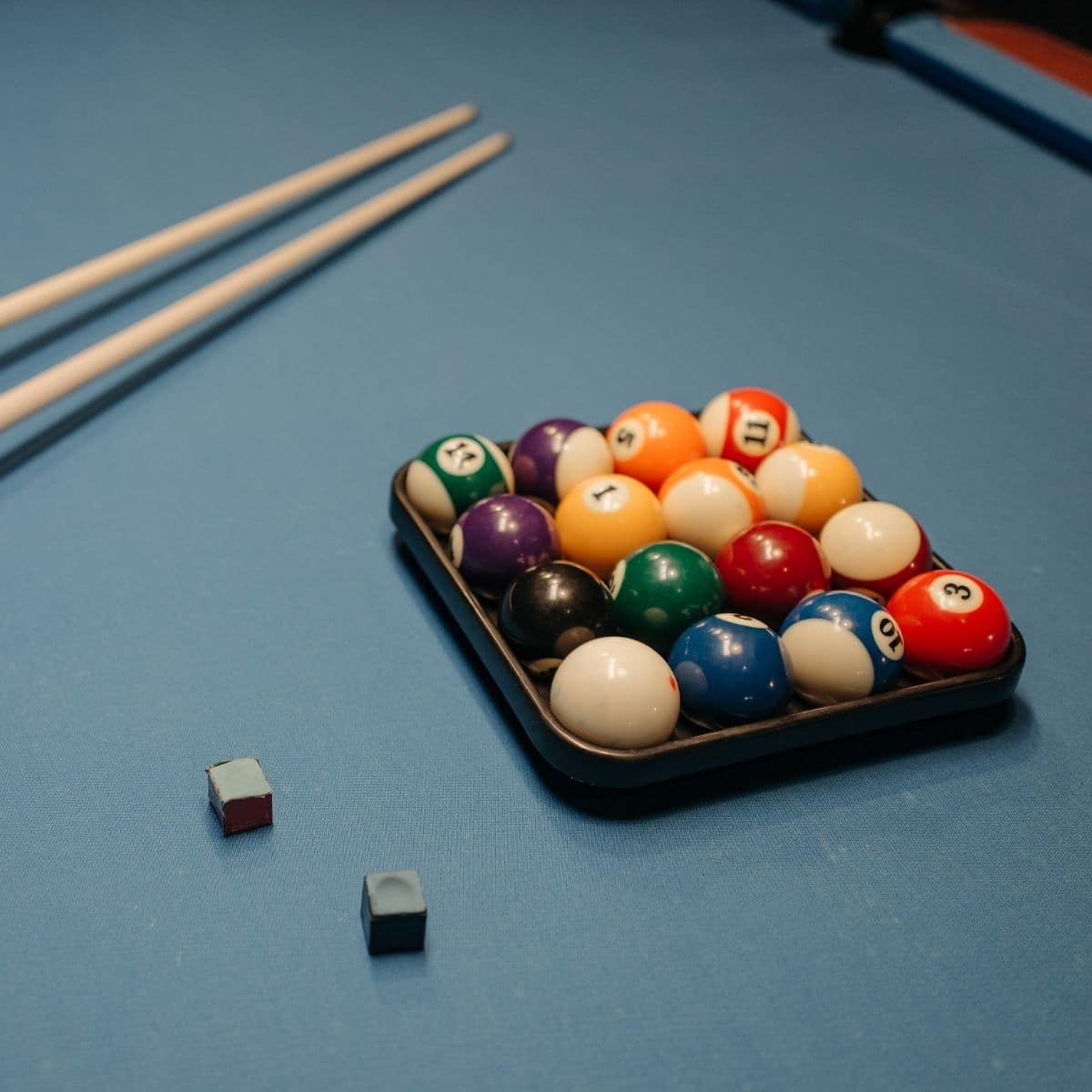 A set of billiard balls arranged in a triangle rack on a blue snooker table, with two cues and two chalk cubes nearby.