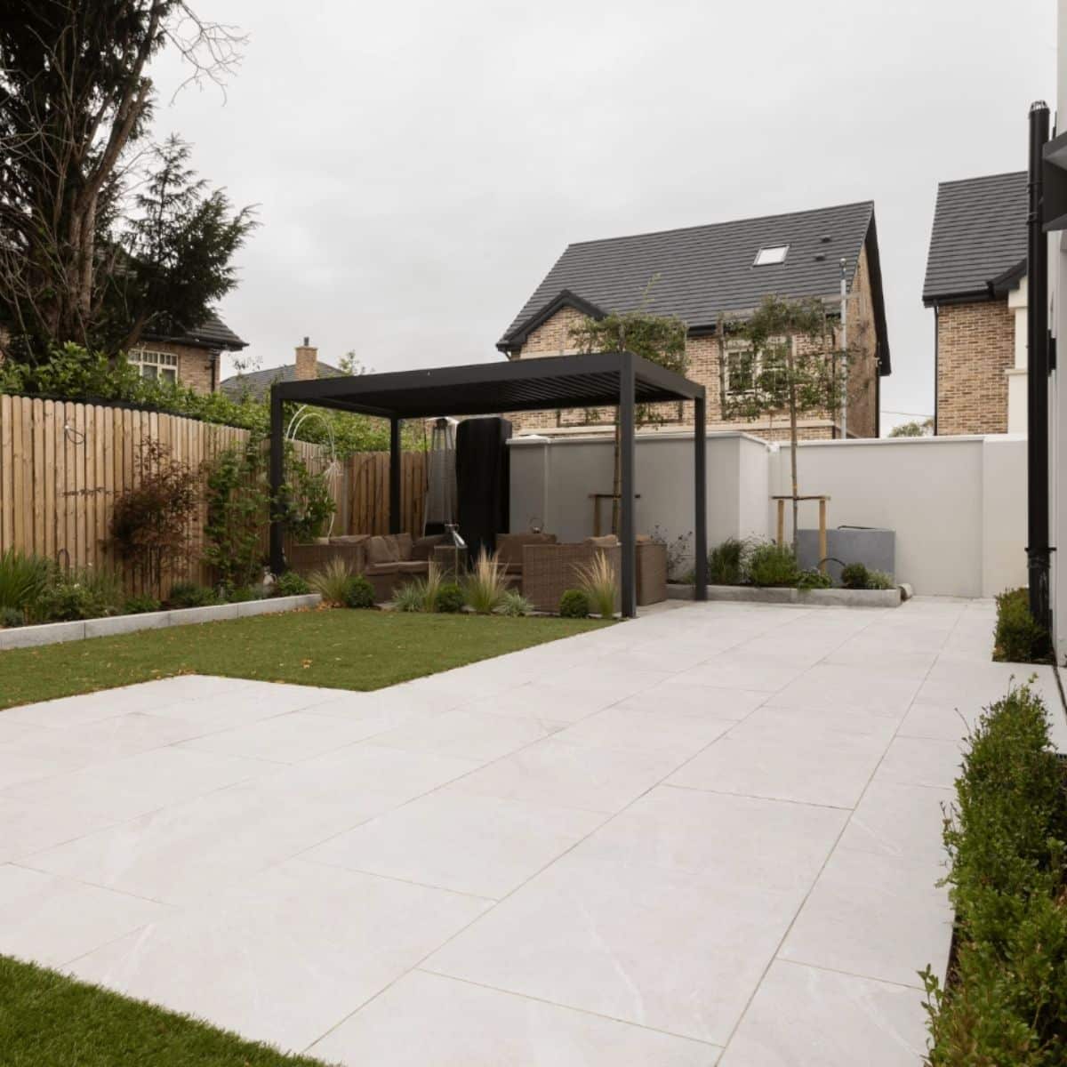 Modern back garden with large white tiles, a grassy area, a pergola with outdoor seating, wooden fence, and neighbouring houses visible in the background under a cloudy sky.