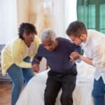 Two carers assist an elderly man in standing up from a bed in a well-lit room with green curtains.
