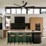 Modern kitchen with wooden cupboards, black appliances, a green island with three wooden stools, large windows, and a ceiling fan overhead.