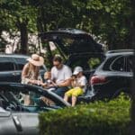 A family of four sits at the open boot of a black SUV parked outdoors, surrounded by other cars and greenery.