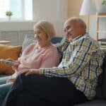 Two older adults sit together on a sofa, smiling and looking towards a television. One holds a remote control. The room has neutral and yellow décor with a lamp and window in the background.