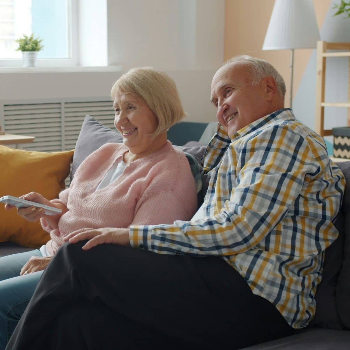 Two older adults sit together on a sofa, smiling and looking towards a television. One holds a remote control. The room has neutral and yellow décor with a lamp and window in the background.