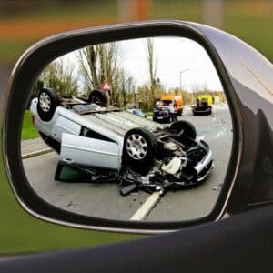 A overturned car with severe damage is seen in a wing mirror, with emergency vehicles and people present at the scene of the accident on the road.