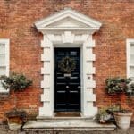 A black door with a wreath, framed by white trim, on a brick house with two potted plants on either side of the entrance.