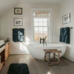 Modern bathroom with a freestanding bath, wooden stool, double towel rails with blue towels, a window with a blind, framed photos on the walls, and wooden flooring.