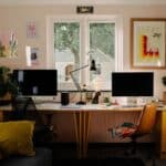 Two desks with computer monitors face a window in a home office; chairs, plants, art prints, and office supplies are arranged neatly around the workspace.