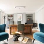 Modern office with a wooden desk, green chair, teal armchairs, wall shelves, and a framed picture. A set of scales and key sit on a round table in the foreground. Room has neutral walls and wood flooring.