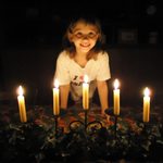 A child stands behind a table with five lit candles arranged in a row, smiling at the camera in a dimly lit room.