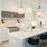 Modern kitchen with white cupboards, marble splashback, stainless steel cooker, large island with sink, two black stools, and two glass pendant lights.
