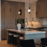Modern kitchen with wooden cupboards, marble splashback, island with sink, two stools, pendant lights, and decorative vases.
