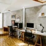 Modern office with two desks, desktop computers, chairs, wall art, shelves, a desk lamp, and wooden flooring. The room has grey textured walls and natural light from a large window.