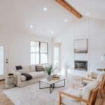 Bright living room with white walls, beige settees, a fireplace, framed art, wooden ceiling beam, large windows, a rug, and a coffee table with a plant.