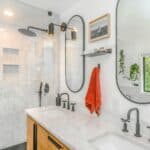 Modern bathroom with marble worktop and shower, double basins, black fittings, an orange towel, oval mirrors, wall shelf with decor, and a hanging plant near a window.