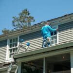 A person in a blue shirt paints an upper window frame on a grey house whilst standing on the roof next to a ladder under a clear sky.