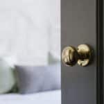 A close-up of a brass door knob on a dark door, partially open, with a blurred bed and pillows visible in the background.